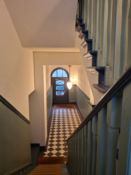 Treppenhaus mit Fliesenboden, aufsteigender Treppe und Türe am Ende des Flurs.